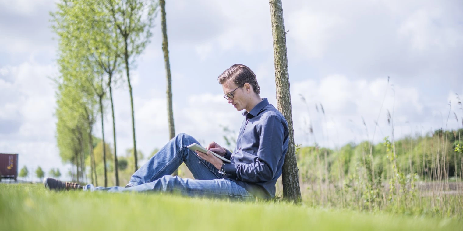 Mann mit Laptop lehnt sitzend an einem Baum und sieht auf sein Tablet